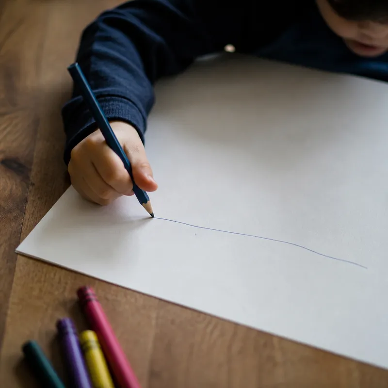 A child's hand draws a single wobbly line across an otherwise blank sheet of paper.
